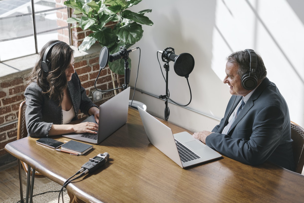 A woman and man sit in front of microphones for a conference call recording in a modern office space.