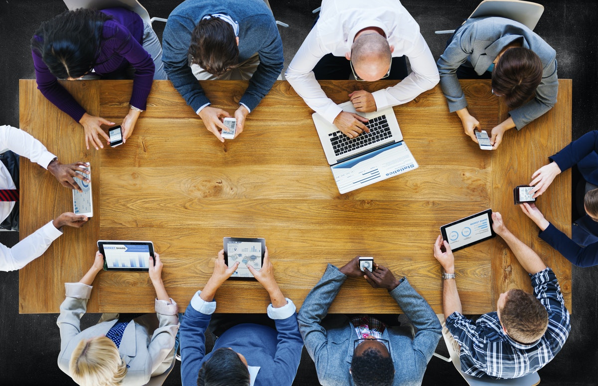 An overhead photo of a work team sitting at a conference table with mobile devices, adopting new technology at their office.